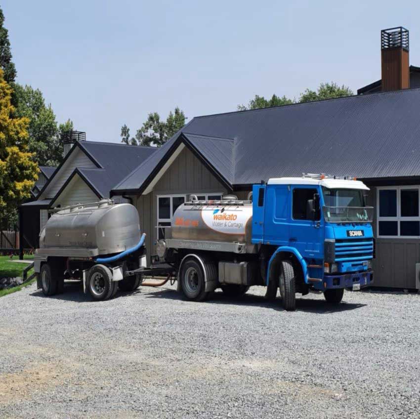 Blue truck with tanker trailer parked in front of a building on a sunny day